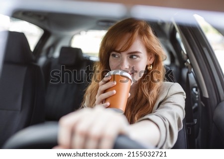 Happy young woman with coffee to go driving her car. Woman sipping a coffee while driving a car. Young woman drinking coffee while driving her car. Attractive red hair drives a car Royalty-Free Stock Photo #2155032721