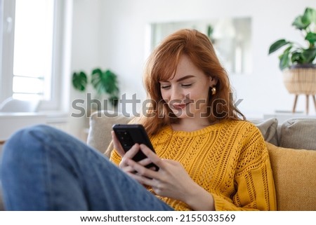 Smiling pretty young woman, relaxes on the sofa in her living room while using her mobile smart phone for social media and surfing the internet
