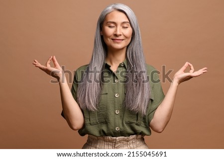 Meditation concept. Calm and satisfied mature woman holds her hands in zen gesture, keeps eyes closed, feels appeasement and pacification, practicing yoga, studio shot isolated on brown