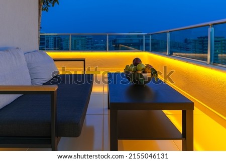View of a cozy outdoor terrace with an armchair, a coffee table and a fruit bowl, LED lighting in the evening. Royalty-Free Stock Photo #2155046131