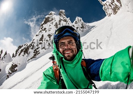 Young smiling skier in helmet, goggles and ski jacket takes a selfie against sun and beautiful snow covered mountain rocks. Male skier makes a photo of himself using his camera.