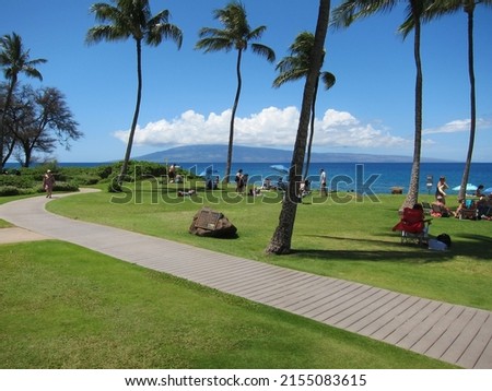 Palm trees, green lawn, clear blue sky, sunny day, people sunbathing and relaxing. View of Lanai island beneath white clouds. Concrete walkway across. Kaanapali, Kahekili Beach Park, Maui, Hawaii. Royalty-Free Stock Photo #2155083615
