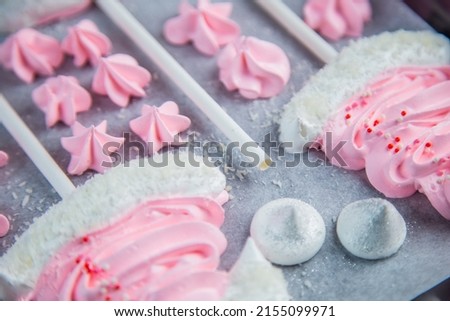 Meringues in the form of pink Christmas hats with a pompom and a white lapel on a stick lie on white parchment in two rows, pink stars and white meringues lie between them. High quality photo