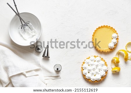 Mini lemon pies with meringue, lemon peel, whisks in meringue, piping nozzles and a white napkin, on a white background with copy space. 