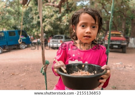 Latin girl sitting on a swing eating from a plastic bowl. Concept of childhood, nutrition and food in rural areas of Nicaragua Latin America Royalty-Free Stock Photo #2155136853