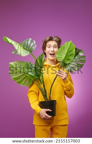 A happy smiling boy teenager in bright yellow hoodie smiles joyfully holding a room plant. Young people, lifestyle. Education, biology and environment.
