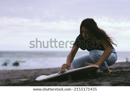Young woman surfer, preparing a surfboard on the ocean, waxing. Woman with safboard on the ocean, active lifestyle, water sports.
