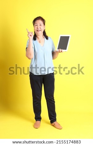 The Asian senior woman wearing light blue shirt standing on the yellow background.