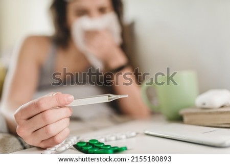 A young woman lies in bed and blows her nose into a handkerchief, takes her temperature and reaches for the pills with her hand. Self-isolation and sick leave at home. Cold Royalty-Free Stock Photo #2155180893