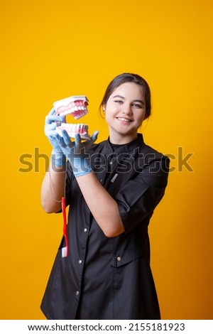 A young girl doctor in black medical uniform holds a mould of jaws and toothbrush on a yellow background