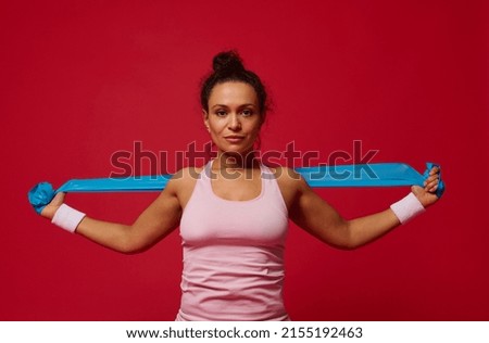 Beautiful confident motivated sportswoman working out with blue elastic band on red background and looking at camera. Copy ad space