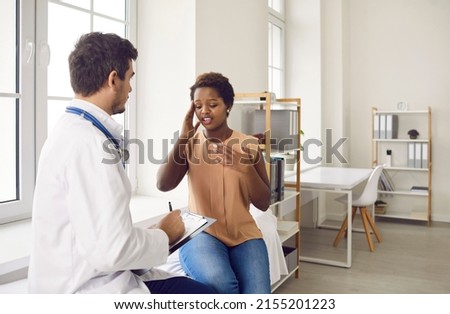 Embarrassed young dark-skinned woman tells doctor about headache during visit to clinic. Sick woman sits on examination couch and provides information about herself to doctor who writes on clipboard. Royalty-Free Stock Photo #2155201223