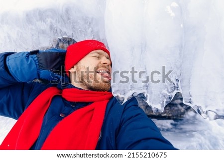 Adventure travel in winter, tourist man with red scarf makes selfie background ice grotto and cave, frozen icicles of Lake Baikal.