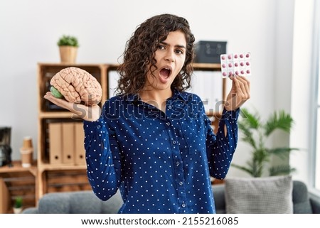 Young brunette woman holding brain and pills as mental health concept in shock face, looking skeptical and sarcastic, surprised with open mouth 