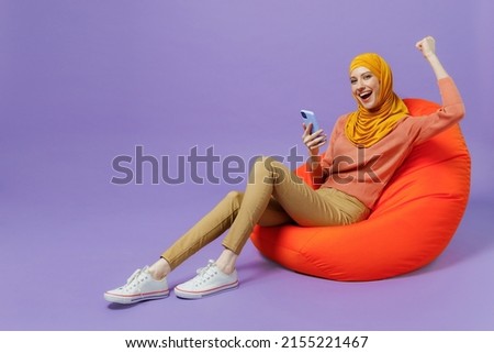 Full size young arabian asian muslim woman in abaya hijab yellow clothes sit in bag chair old use mobile cell phone do winner gesture isolated on plain pastel light violet background studio portrait