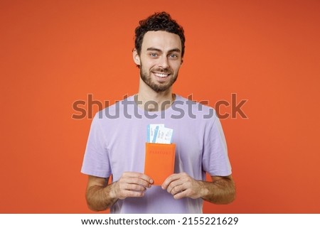 Young man 20s in violet t-shirt hold passport tickets looking camera isolated on orange background studio. Passenger travel abroad on weekends getaway. Air flight journey concept. Tattoo translate fun