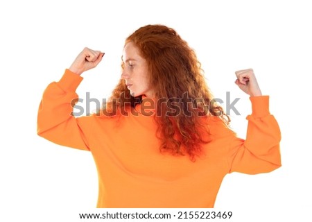 Smiling young redhead girl wearing orange sweater over white background raises hand to show muscles, feels confident in victory, strong and independent. 