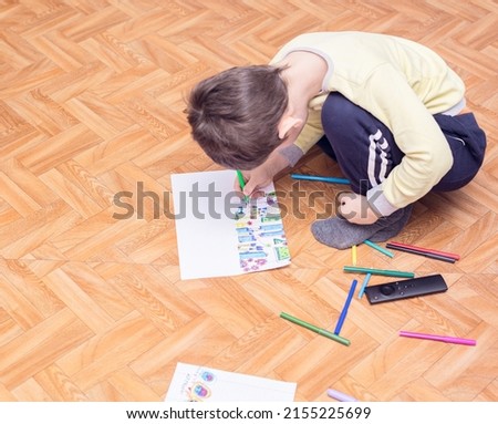 The child boy sits on the floor and draws a picture with colored markers on white sheet. Leisure activity at home