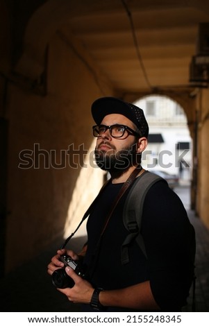 Street portrait of a male photographer with a beard in glasses and a cap with a vintage camera. Young hipster man with digital vintage camera and backpack looks up.