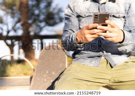 Adult man texting his friends on his phone sitting with his skateboard next to him.
