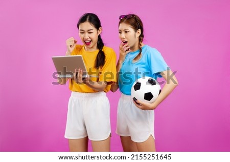 Young asian women in yellow and blue t-shirt holding ball looking and cheering a world cup soccer football game on tablet pink screen background.