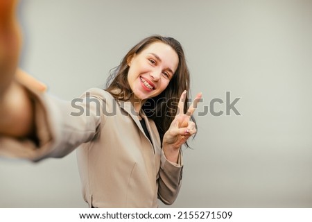 Smiling lady in beige jacket taking selfie on camera smartphone on beige background, looking at camera with happy face.