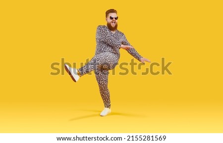 Plus size guy in funny pyjamas having fun in modern studio. Happy carefree fat man wearing comfortable leopard PJs and sunglasses dancing isolated on yellow background. Crazy party and fashion concept Royalty-Free Stock Photo #2155281569