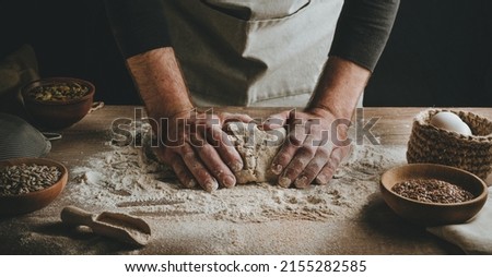 Unrecognizable young man kneading dough on wooden table. Males hands making bread on dark background. Selective focus. Royalty-Free Stock Photo #2155282585