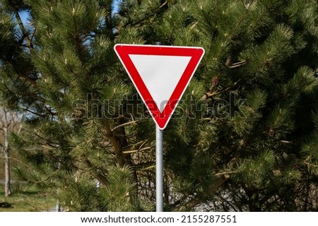 Give way road sign next to crossroads. Also called yield sign to guide traffic. Triangular shape with a red stroke. Warning symbol to beware of the traffic crossing the way.