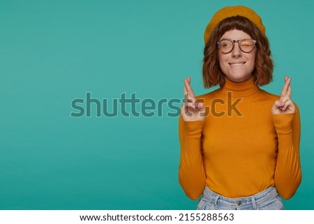 Indoor portrait of young ginger female, wears orange sweatshirt and spectacles posing over blue background keeps her fingers crossed and looks aside while praying for good exams result