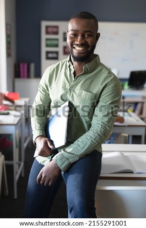 Portrait of smiling african american young male teacher with file sitting at desk in classroom. unaltered, education, teaching, occupation and school concept. Royalty-Free Stock Photo #2155291001