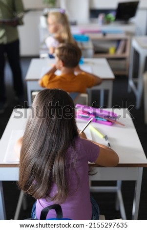 Rear view of caucasian elementary schoolgirl writing on book at desk in classroom. unaltered, education, learning, childhood, teaching and school concept.