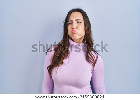 Young brunette woman standing over blue background puffing cheeks with funny face. mouth inflated with air,  expression. 