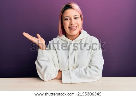 Hispanic woman with pink hair wearing casual sweatshirt sitting on the table smiling cheerful presenting and pointing with palm of hand looking at the camera. 