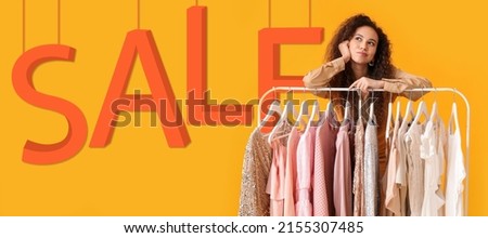 Beautiful thoughtful African-American girl near rack with clothes and word SALE on yellow background