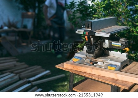 circular saw in the foreground located in the garden with man in work bib behind it working