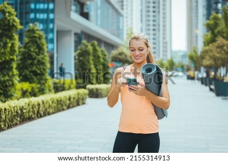 Sporty woman listening to music over the wireless earphones in an urban area. Sporty fit woman poses with sport equipment, uses smartphone for tracking fitness results dressed in sportswear 