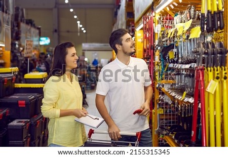 Happy married couple chooses variety of tools and materials for repairs in apartment. Young husband and wife are looking at goods on shelves of large warehouse hypermarket of building materials. Royalty-Free Stock Photo #2155315345