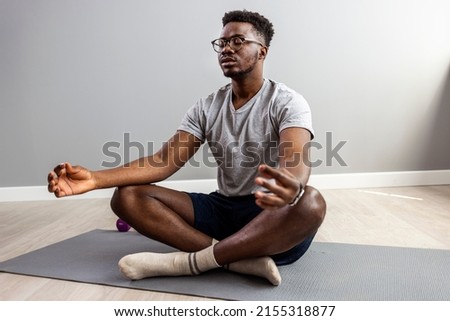 Man in sporty outfit doing yoga and meditating on an exercise mat. Sporty mindful man with tattoo meditating alone at home, peaceful calm hipster fit guy practicing yoga in lotus pose  Royalty-Free Stock Photo #2155318877