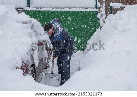 A man is clearing the street of snow with a shovel. Nearby is a red car covered with snow. Large flakes of snow falling. Cleaning the street during a snow storm Royalty-Free Stock Photo #2155321173
