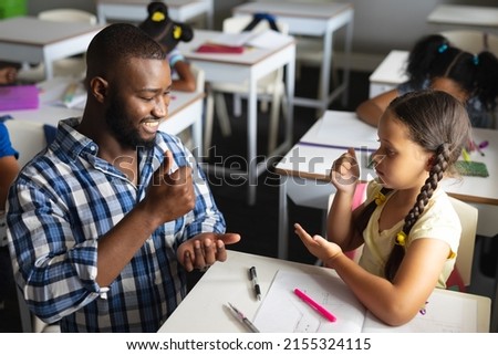 African american young male teacher teaching sign language to caucasian elementary girl in class. unaltered, education, childhood, learning, teaching, disability and school concept.