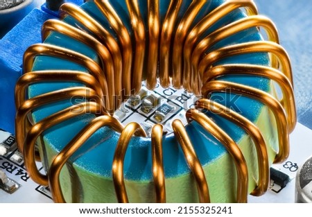 Close-up of ferrite core coil with copper wire and resistors or capacitors on PCB detail. Toroidal electronic inductor metal winding. White circuit board with surface mount devices on blue background. Royalty-Free Stock Photo #2155325241