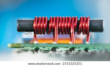 Electronic transformer with ferromagnetic core and coil copper wire on PCB. Closeup of red cylindric inductor soldered on green circuit board with tin on bottom side and blue background. Cable TV amp. Royalty-Free Stock Photo #2155325251