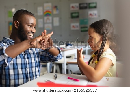Smiling african american young male teacher teaching sign language to caucasian elementary girl. unaltered, education, childhood, learning, teaching, disability and school concept. Royalty-Free Stock Photo #2155325339