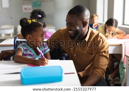 Smiling african american young male teacher looking at african american elementary schoolgirl. unaltered, education, childhood, disability, learning, physical disability and school concept. Royalty-Free Stock Photo #2155327977