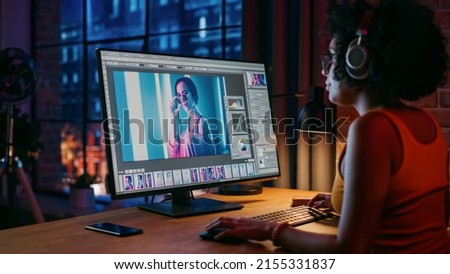 Young Creative Female in Headphones Using Computer in Stylish Loft Apartment in the Evening. Graphic Designer Working from Home, Editing Fashion Photo for Online Store. Urban View from Big Window.