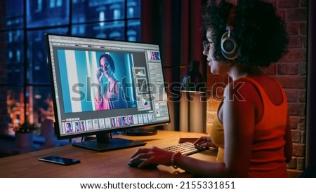 Young Creative Female in Headphones Using Computer in Stylish Loft Apartment in the Evening. Graphic Designer Working from Home, Editing Fashion Photo for Online Store. Urban View from Big Window.