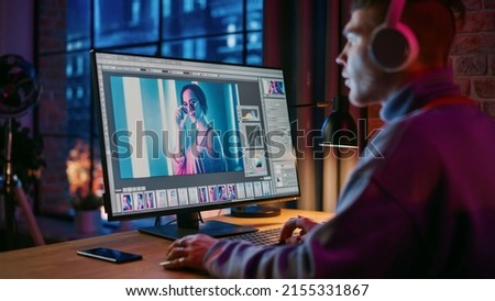 Young Creative Man in Headphones Using Computer in Stylish Loft Apartment in the Evening. Graphic Designer Working from Home, Editing Fashion Photo for Online Store. Urban City View from Big Window.