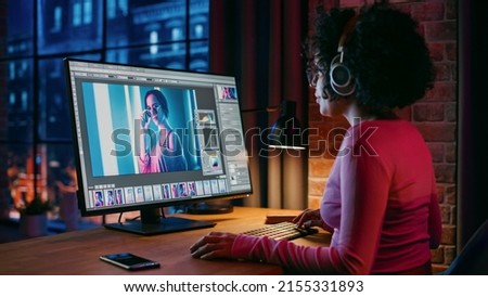 Young Creative Female in Headphones Using Computer in Stylish Loft Apartment in the Evening. Graphic Designer Working from Home, Editing Fashion Photo for Online Store. Urban View from Big Window. Royalty-Free Stock Photo #2155331893