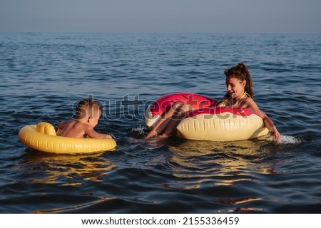 A young mother and a child in swimming circles have fun and laugh while swimming in the sea.